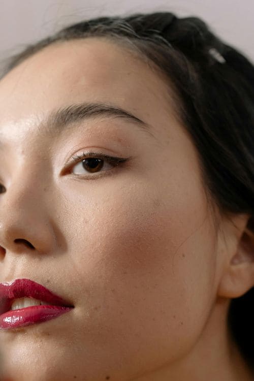 Une femme tient un miroir de poche et applique du rouge à lèvres rouge vif sur ses lèvres. Elle est bien éclairée, et l’image est centrée sur son visage et sa main. L’ambiance est intimiste et élégante, avec une mise au point sur le geste de maquillage.