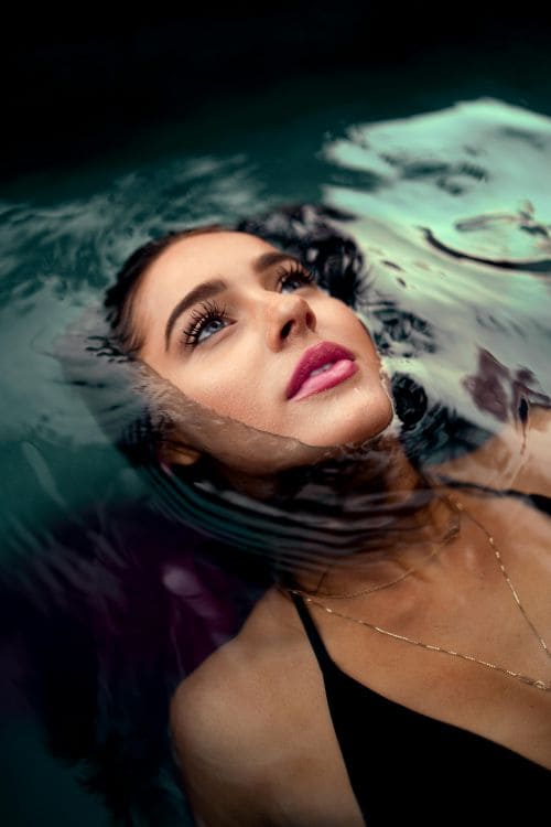 Une femme flotte paisiblement à la surface de l’eau, le visage partiellement immergé, les yeux ouverts et regardant vers le ciel. Elle porte un maillot noir et un collier fin en or.</p>
<p>